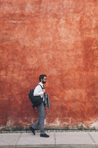Side view of businessman with backpack walking on sidewalk against wall in city