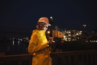 Female professional wearing yellow raincoat and hardhat using digital tablet at night