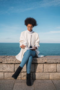 Young woman using digital tablet while sitting on retaining wall against sea