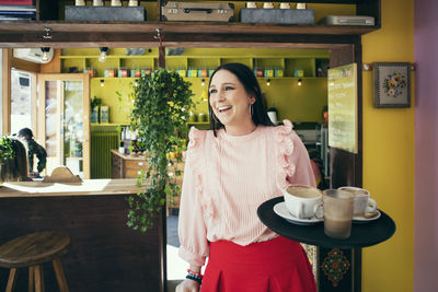 Happy waitress holding serving tray while looking away in cafe