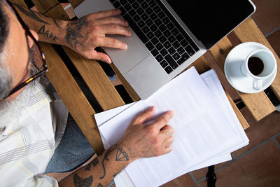 From above of crop unrecognizable male in casual clothes with tattooed arms browsing laptop while working remotely at table in home workplace