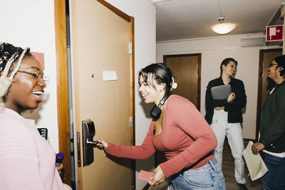 Happy young woman opening door while multiracial friends at corridor in college dorm