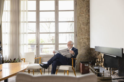 Senior man reading newspaper sitting by window in living room