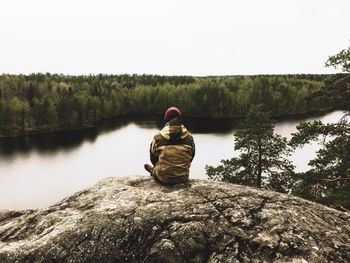 Rear view of man sitting on rock by river in forest against clear sky