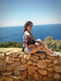 Full length of young woman sitting on retaining wall by sea