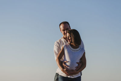 Smiling man touching pregnant girlfriend belly while standing against clear sky