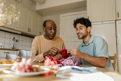 Smiling male caregiver knitting with senior male sitting in kitchen at home