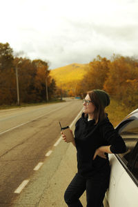 Girl in swamp hat and hoodie drinking coffee in craft glass on the side of an autumn forest road