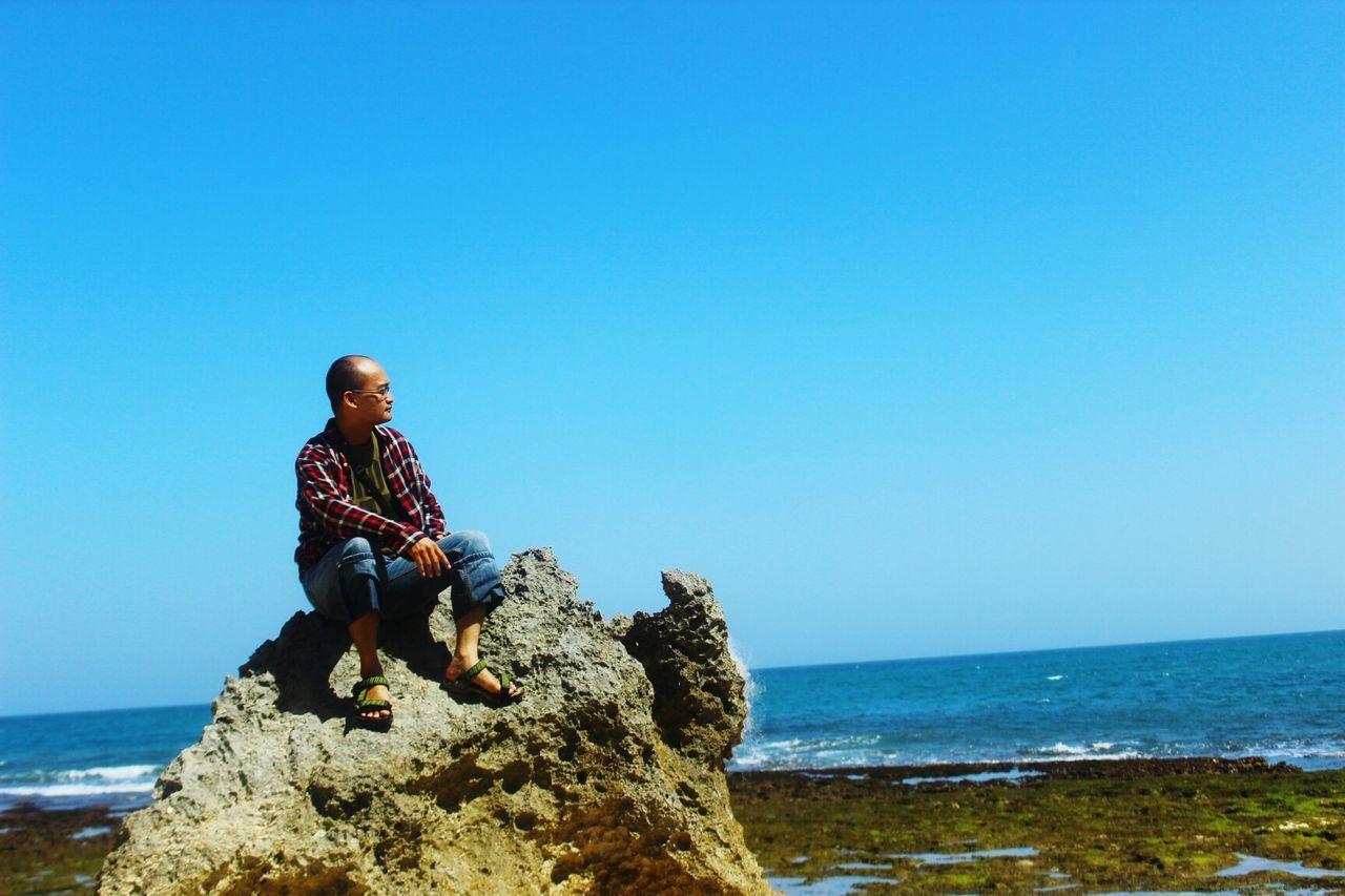 sea, horizon over water, clear sky, copy space, beach, water, lifestyles, leisure activity, blue, shore, rock - object, scenics, men, beauty in nature, vacations, tranquility, full length, sitting