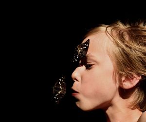 Close-up of boy with butterfly perching on face against black background