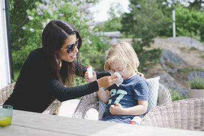 Mother assisting son in blowing nose while sitting on chair at yard