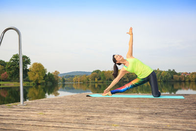 Smiling woman exercising on pier over river against sky