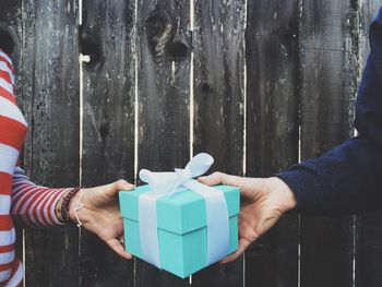 Cropped image of hands holding gift box against wooden fence