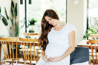 Caucasian young redhead freckled pregnant woman with long hair in a white dress hugs her belly. 