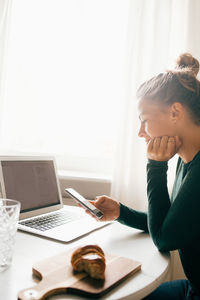 Mid adult woman using smart phone while surfing food recipe on laptop at home