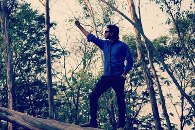 Low angle view of young man with hand raised standing on tree trunk in forest