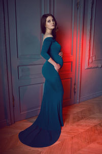 Pregnant woman in a long blue dress standing against the wall in the studio
