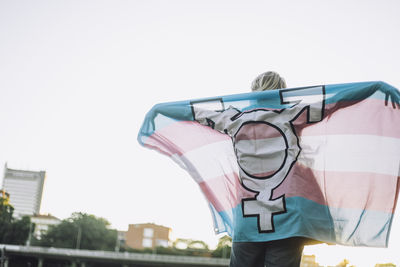 Rear view of non-binary person standing with lgbtqia flag against clear sky