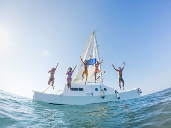 Low angle view of playful friends jumping from boat in sea against clear blue sky