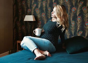 Pregnant woman sitting on bed while holding cup in bedroom