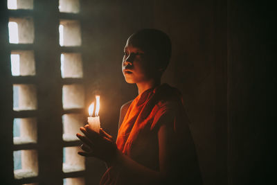 Young woman looking away while standing against window in dark room