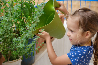 Little girl waters greenery with a watering can at balcony. child helps to take care about plants