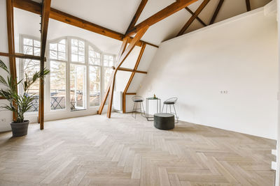 Small square table with chairs and pouf placed in spacious room with wooden floor and beams decorated with potted plant in daylight