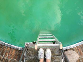 Low section of person standing by ladder in turquoise pool