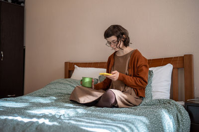 Teen girl reading messages scrolling social media in smartphone drinking tea in bed.