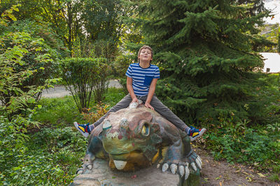 Cheerful smiling boy in a striped t-shirt sits on a dinosaur statue in the park at the playground