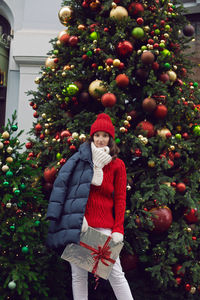 Young woman in a red knitted hat and sweater stands with a gift at the christmas tree