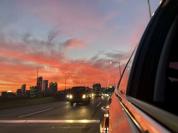 Cars on road by buildings against sky during sunset