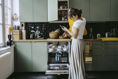 Smiling woman sharing smart phone to boyfriend doing dishes in kitchen at home