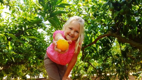 Portrait of happy girl giving orange by trees