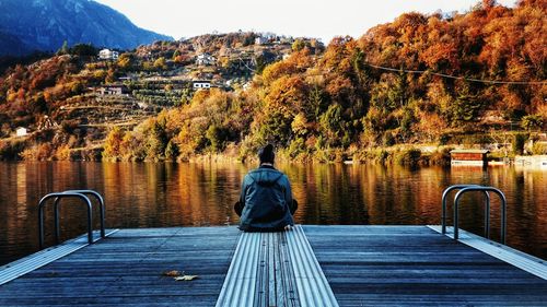 Rear view of man sitting on jetty over lake against trees