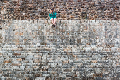 Full length of woman photographing while sitting on brick wall