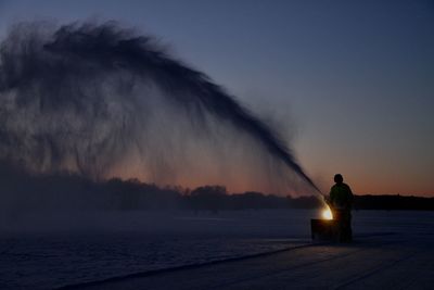 Man using snowblower on frozen lake during sun set