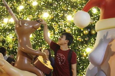 Young man touching reindeer balloon against illuminated christmas tree at night