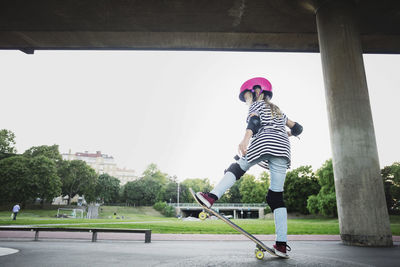 Full length of girl performing stunt with skateboard at park against clear sky