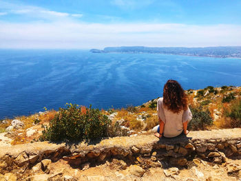 Rear view of woman sitting on rock by sea against sky