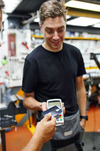 Crop customer putting credit card over contactless terminal to pay to mechanic for repair service in modern garage