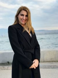 Portrait of smiling young woman standing at beach against sky