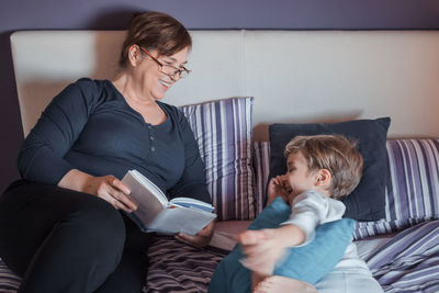 Grandmother reading book to grandson while relaxing on bed at home