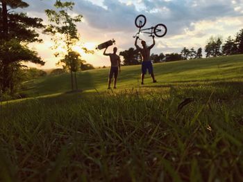 Full length of men standing with skateboard and bicycle on grass at park during sunset