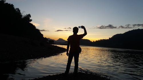 Rear view of silhouette man standing by lake against sky during sunset
