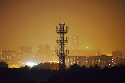 Silhouette communication tower against illuminated buildings and sky at dusk