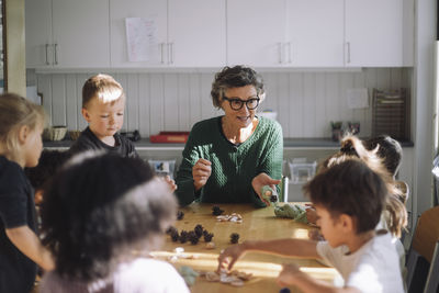 Senior female teacher explaining clay molding to preschool kids sitting in classroom at kindergarten