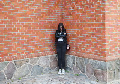 Young beautiful brunette girl in black jacket and jeans stands near wall of gothic church on summer