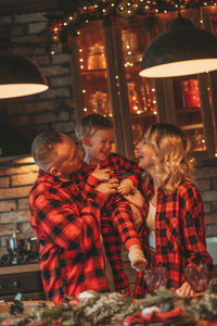 Smile active parents with small son in red checkered sleepwears waiting santa indoor.