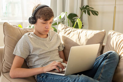 Portrait of teenage , sits on a sofa , wearing black headphones on his head, using a laptop.